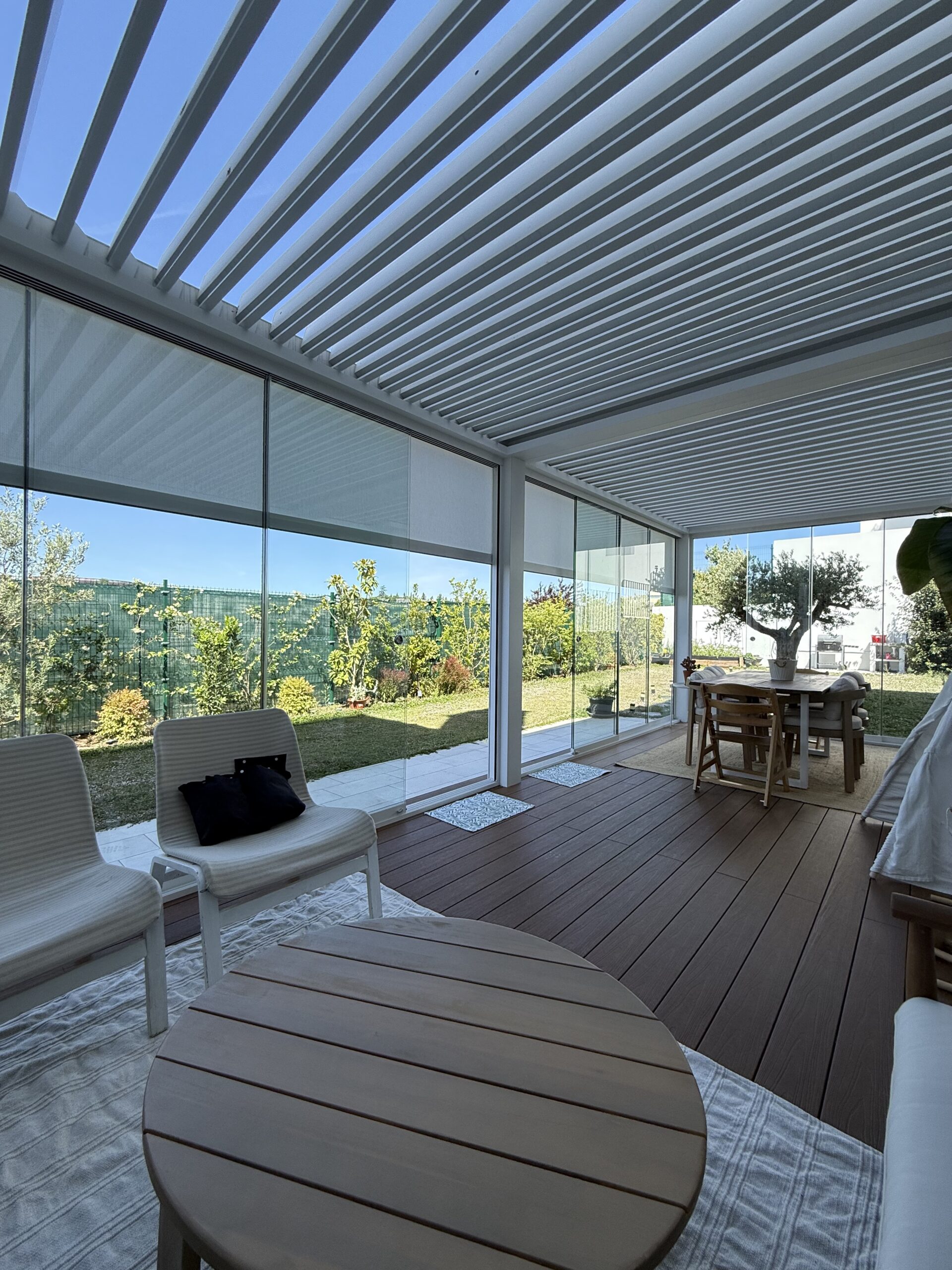 Salón exterior cubierto con cortinas de cristal correderas, sillones y alfombra junto al jardín de una vivienda unifamiliar en Madrid · Two Brothers Madrid.
